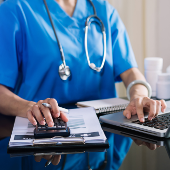 Medical office professional in scrubs using a laptop and calculator to manage paperwork and administrative tasks in a healthcare setting.