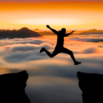 A Silhoutte of a man jumping over a gap, with cloudy mountains and a sunrise in the background.
